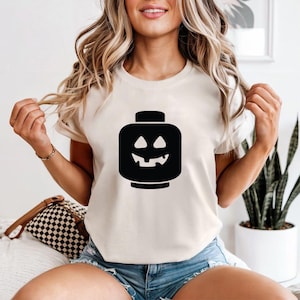May include: A cream-colored t-shirt featuring a black jack-o'-lantern design. The graphic is a stylized Lego head with a carved pumpkin face. The shirt has short sleeves and a crew neck.