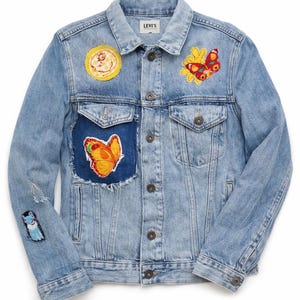 May include: Light blue denim jacket with embroidered patches. The patches include a sun, butterflies, and a dog. The jacket has a button-up front, two chest pockets, and the Levi's logo on the label.