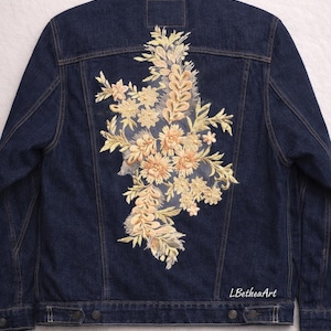 May include: Dark blue denim jacket with an embroidered floral design on the back. The embroidery features beige and light orange flowers and leaves. The text "LBetheaArt" is visible on the lower right side of the jacket.