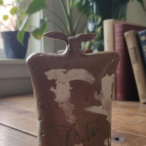 Op de afbeelding: Een bruine keramische vaas met een unieke vorm en een handvat. De vaas heeft witte en groene accenten, met het woord "LYONS" ingeschreven. Hij staat op een houten oppervlak.