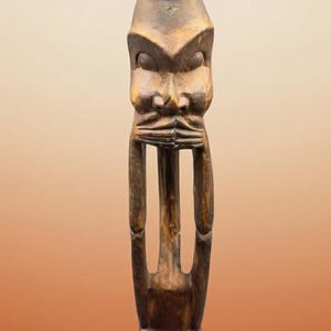 May include: A carved wooden figure with a large head and a small pot on top. The figure has a stylized face and arms, and it stands on a square base. The wood is a dark brown colour.