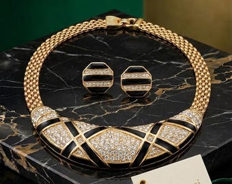 May include: A gold-toned necklace and matching earrings set. The necklace features a black and crystal design, with a mesh chain. The earrings have a black and crystal design. A tag with the text "Nina Ricci" is included.