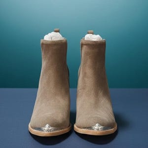 May include: A pair of taupe suede ankle boots with silver-toned metal accents on the toes. The boots have a stacked heel and are stuffed with white paper. The background is a gradient of teal and navy blue.