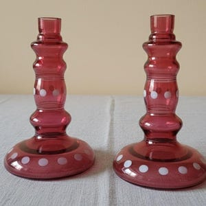 Może przedstawiać: Dwa pasujące świeczniki z rubinowego szkła. Każdy ma okrągłą podstawę, pękaty środek i górną część do trzymania świecy. Białe grochy zdobią korpus każdego świecznika. Świeczniki mają około 15 cm wysokości.