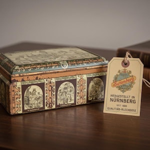 May include: A vintage rectangular tin box with detailed illustrations and a distressed finish. A tan tag with the text "Buremberg" and "Nürnberg" hangs nearby. A collectible item, possibly from Germany.
