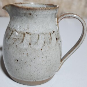 May include: A handmade ceramic jug with a light grey glaze and brown speckles. The jug has a curved handle and a spout for pouring. The base and rim are a darker brown. The jug has a textured surface and a decorative pattern.