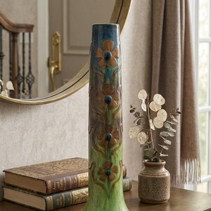 May include: A decorative ceramic vase with a floral design in green, brown, and blue. The vase is on a wooden table with books, a brass dish, and a small vase with dried flowers. The table is near a mirror and a brown leather briefcase.