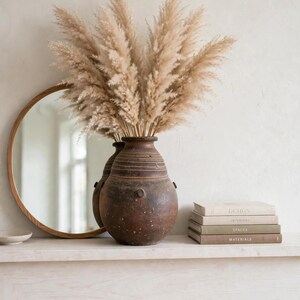 Puede incluir: Un jarrón de cerámica marrón oscuro, redondo y texturizado, lleno de altos y esponjosos penachos de hierba de la pampa beige. Junto al jarrón hay una pila de cuatro libros titulados Design, Interiors, Spaces y Materials, y un espejo redondo.