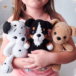 May include: Three plush crochet stuffed animals including a white dog with black spots, a black, white, and brown tri-color dog, and a solid tan dog. Each toy features black plastic eyes, a black nose, and soft yarn texture. Text reads CrochetPolyaToys.