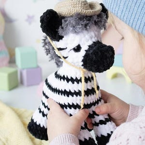 Op de afbeelding: Een handgemaakt gehaakt zebra knuffel met zwart-witte strepen, een zwarte neus en een kleine beige hoed. De zebra heeft een gele draad om zijn nek. Het speelgoed wordt vastgehouden door een persoon die een roze trui draagt.