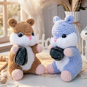 Puede incluir: Dos peluches de hámster hechos a mano con ganchillo. Uno es marrón y blanco, el otro azul y blanco. Ambos tienen patas y narices rosas y sostienen un objeto gris oscuro. Los juguetes están sobre una superficie blanca.