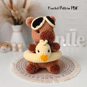May include: A brown crocheted bear plush toy wearing black and white sunglasses, holding a small yellow crocheted duckling inside a pale yellow crocheted swim ring. The text Crochet Pattern PDF is visible at the top of the image.