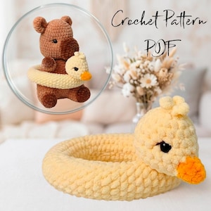 May include: A crochet pattern PDF featuring a brown bear plush toy wearing a yellow duck-shaped swim ring. The image also shows a separate yellow duck-shaped swim ring made of soft, textured yarn with a bright orange beak and black eyes.