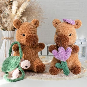 May include: Two brown crochet bears with dark brown snouts and small black eyes. One bear holds a green turtle bag with a pink head, and the other holds a purple flower. A small purple flower is on the bear's head.