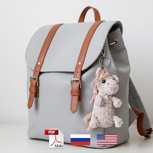 Puede incluir: Una mochila gris con correas y hebillas de cuero marrón. Un pequeño adorno de caballo de ganchillo cuelga de la parte delantera. La bolsa tiene un asa superior y correas ajustables. La bolsa está sobre una superficie blanca.