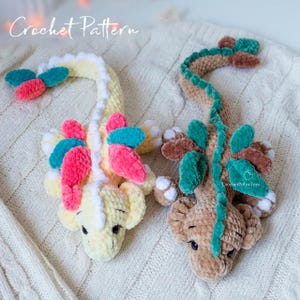 May include: Two plush dragon toys featuring a crochet pattern. One is pale yellow with pink and teal accents, and the other is light brown with dark green accents. Both have white details on their feet and tails, and black eyes. Text reads Crochet Pattern.