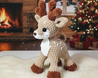May include: A crocheted reindeer toy with brown antlers, hooves, and a white chest. The reindeer has large black eyes and is standing on a white surface. A Christmas tree and fireplace are visible in the background.