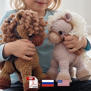 May include: Two handmade crochet horse plush toys. One is brown with a dark brown mane and the other is light pink with a white mane. The toys are held by a person. The toys are soft and cuddly.