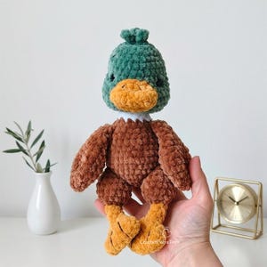Puede incluir: Un juguete de pato tejido a ganchillo con cabeza verde oscuro, pico naranja brillante, cuello blanco y cuerpo marrón con pies naranjas. El juguete tiene un acabado de hilo suave y texturizado e incluye el texto CrochetPolyaToys en el cuerpo.