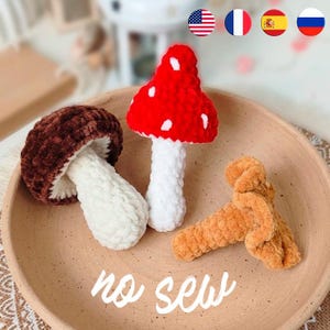 May include: Three crocheted mushroom figures in a bowl. One has a dark brown cap and white stem, one has a bright red cap with white spots and a white stem, and one is a solid light brown color. The text "no sew" is written in white script.