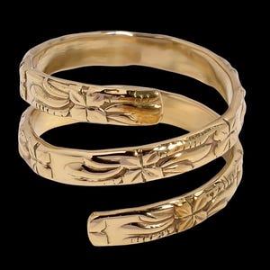May include: A gold-colored, spiral ring with an engraved floral pattern. The ring wraps around the finger in a three-turn design. The ring is made of a shiny, metallic material and is set against a black background.