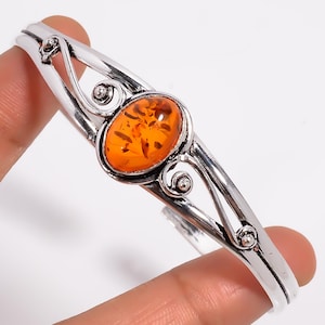May include: A silver cuff bracelet with an oval amber stone. The amber is a warm orange colour with visible inclusions. The bracelet has decorative swirls and a polished finish, suitable for everyday wear.