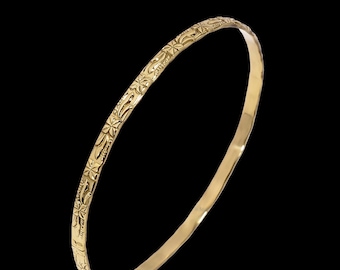 May include: A gold-coloured bangle bracelet with an intricate floral pattern. The bracelet is circular and appears to be made of metal. The design features repeating star-like shapes and other decorative elements. The background is black.