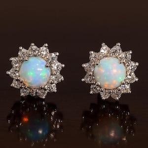 May include: A pair of opal stud earrings. Each earring features a round, iridescent opal stone surrounded by a halo of small, clear gemstones. The earrings are set in a silver-colored metal, creating a sparkling, elegant design.