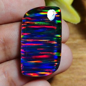 Op de afbeelding: Een rechthoekige zwarte opaal edelsteen met levendige horizontale banden van rood, blauw, groen en geel. De gepolijste steen heeft een afgeronde bovenrand en wordt in een hand gehouden, wat zijn kleurrijke kleurenspel laat zien.