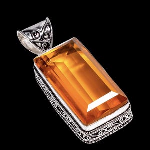 May include: A rectangular pendant featuring a large, faceted, amber-colored gemstone set in a decorative silver-toned metal frame. The pendant has an ornate bail with a swirling design, and the gemstone is a warm, golden hue.