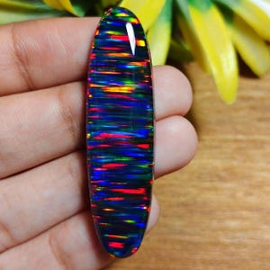 May include: A polished, elongated black opal gemstone with vibrant streaks of red, green, and yellow. The opal is held in a hand, showcasing its colorful play-of-color. The stone's shape is oval, and it appears to be a cabochon cut.