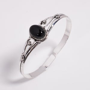 May include: Silver bangle bracelet with an oval, black gemstone. The bracelet has decorative swirls and a polished finish. A piece of jewellery suitable for everyday wear or special occasions.
