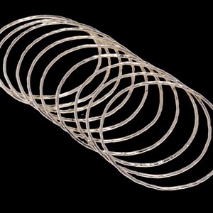 Peut inclure: Ensemble de bracelets argentés avec une finition texturée et martelée. Les bracelets sont disposés en chevauchement léger sur fond noir, créant un effet de superposition. Les bijoux sont simples et élégants.