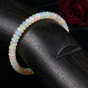 Könnte beinhalten: Ein Armband aus Opalperlen mit schillernden Farben. Das Armband besteht aus kleinen, runden Opalperlen, die aneinandergereiht sind. Die Perlen zeigen eine Reihe von Farben, darunter Weiß, Grün und Orange. Das Armband wird auf einer dunklen Holzoberfläche präsentiert.