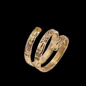 May include: A gold-colored, spiral ring with floral engravings. The ring wraps around in three loops, with detailed carvings along its surface. The ring is set against a black background, highlighting its intricate design and metallic sheen.