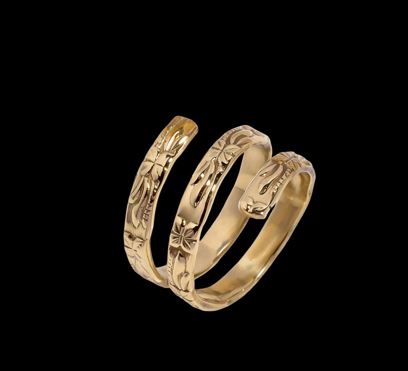 May include: A gold-colored, spiral ring with floral engravings. The ring wraps around in three loops, with detailed carvings along its surface. The ring is set against a black background, highlighting its intricate design and metallic sheen.