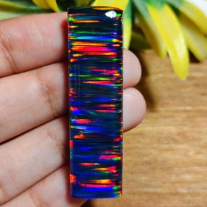 Puede incluir: Una piedra preciosa de ópalo negro rectangular y pulida con un vibrante fuego interno. El ópalo muestra bandas de rojo, naranja, amarillo, verde y azul, creando un llamativo juego de colores. La piedra preciosa se sostiene en una mano.