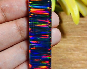 May include: A rectangular, polished black opal gemstone with vibrant internal fire. The opal displays bands of red, orange, yellow, green, and blue, creating a striking play of color. The gemstone is held in a hand, showcasing its polished surface.