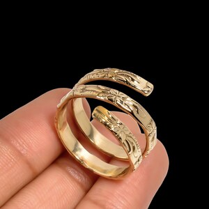 May include: A gold-colored spiral ring with floral engravings. The ring wraps around the finger with a three-band design. The ring is held against a black background.