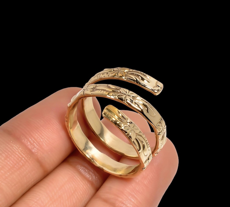 May include: A gold-colored spiral ring with floral engravings. The ring wraps around the finger with a three-band design. The ring is held against a black background.