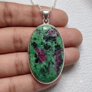 May include: An oval-shaped pendant featuring a polished ruby zoisite gemstone with a mottled pattern of vibrant green, deep magenta, and black inclusions. The stone is set in a simple silver-toned metal bezel and hangs from a silver-toned chain.