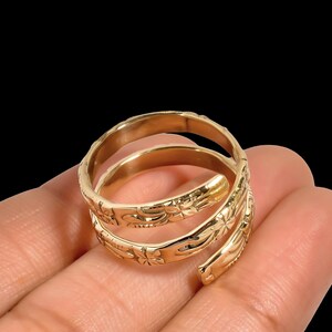 May include: A gold-colored spiral ring with floral and leaf patterns. The ring wraps around the finger in a three-turn design. The ring is held in a hand against a black background.