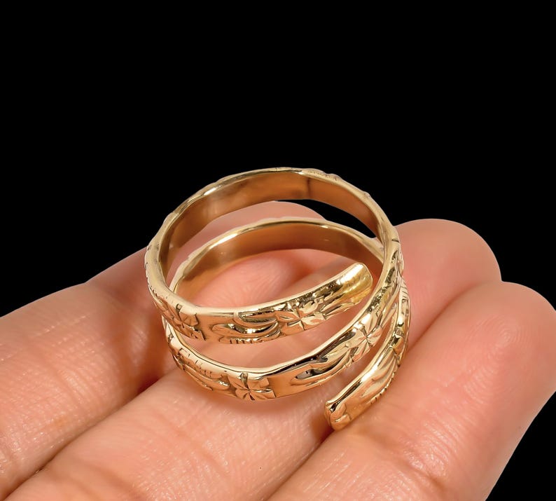 May include: A gold-colored spiral ring with floral and leaf patterns. The ring wraps around the finger in a three-turn design. The ring is held in a hand against a black background.
