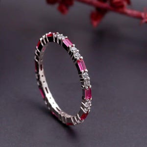 May include: A silver ring with alternating red and clear stones. The ring features rectangular red stones and small, round clear stones set in a continuous pattern around the band. The ring is displayed on a dark surface with red floral accents.
