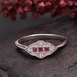 May include: A silver ring with a decorative, rectangular centrepiece. The centre has three square, deep red gemstones, with smaller white stones either side. The ring is set against a dark, textured background, with a blurred floral element in the background.