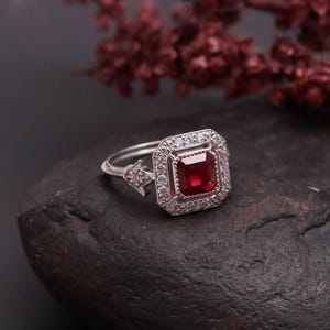 May include: A silver ring with a square, deep red gemstone at the center, surrounded by smaller, clear stones. The ring is set on a dark, textured surface, with a blurred floral element in the background.