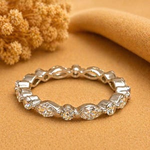 Pode incluir: Um anel prateado com um padrão contínuo de pedras preciosas claras redondas e em forma de marquesa. O anel está sobre um fundo bege, com um elemento floral desfocado e um objeto curvo bege no fundo.