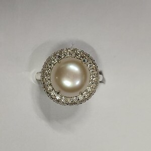 May include: A silver ring featuring a large, lustrous, cream-colored pearl surrounded by a halo of small, sparkling gemstones. The ring's band is thin and silver, and the setting highlights the pearl's round shape.