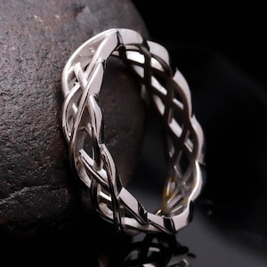 Puede incluir: Un anillo plateado con un diseño de nudo celta. El anillo presenta un intrincado patrón entrelazado, creando un diseño continuo y fluido. El anillo se muestra sobre una superficie oscura y texturizada, resaltando su brillo metálico.