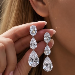 Puede incluir: Un par de pendientes colgantes con tres piedras transparentes facetadas en disposición vertical. La piedra superior es ovalada, mientras que las dos piedras inferiores tienen forma de pera. Cada piedra está engastada en un marco de metal plateado.